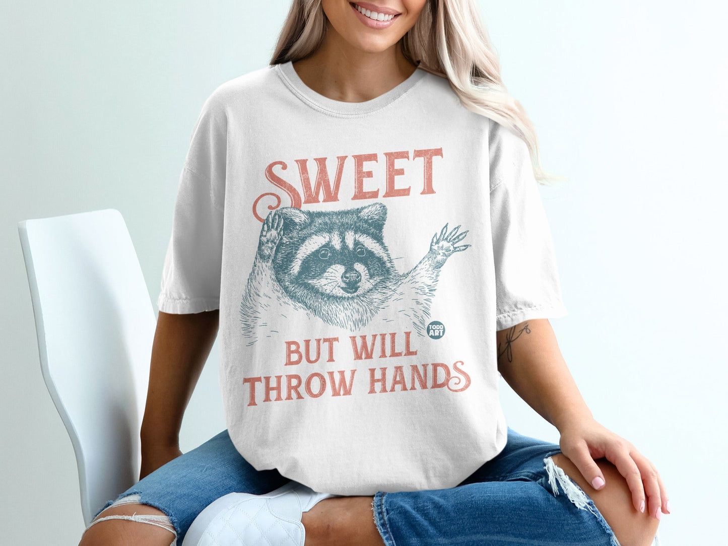 White t-shirt with raccoon illustration and text "SWEET BUT WILL THROW HANDS".