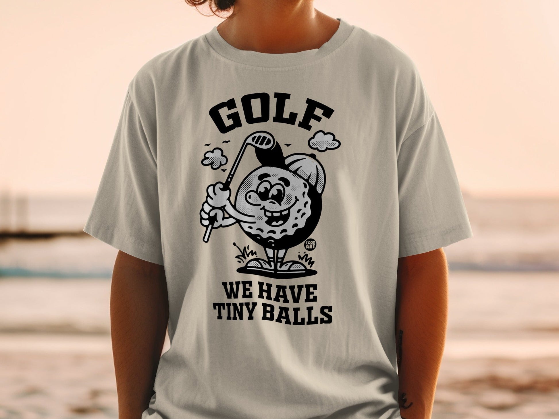 Cartoon golf ball character holding a club, wearing a shirt that says "WE HAVE TINY BALLS".