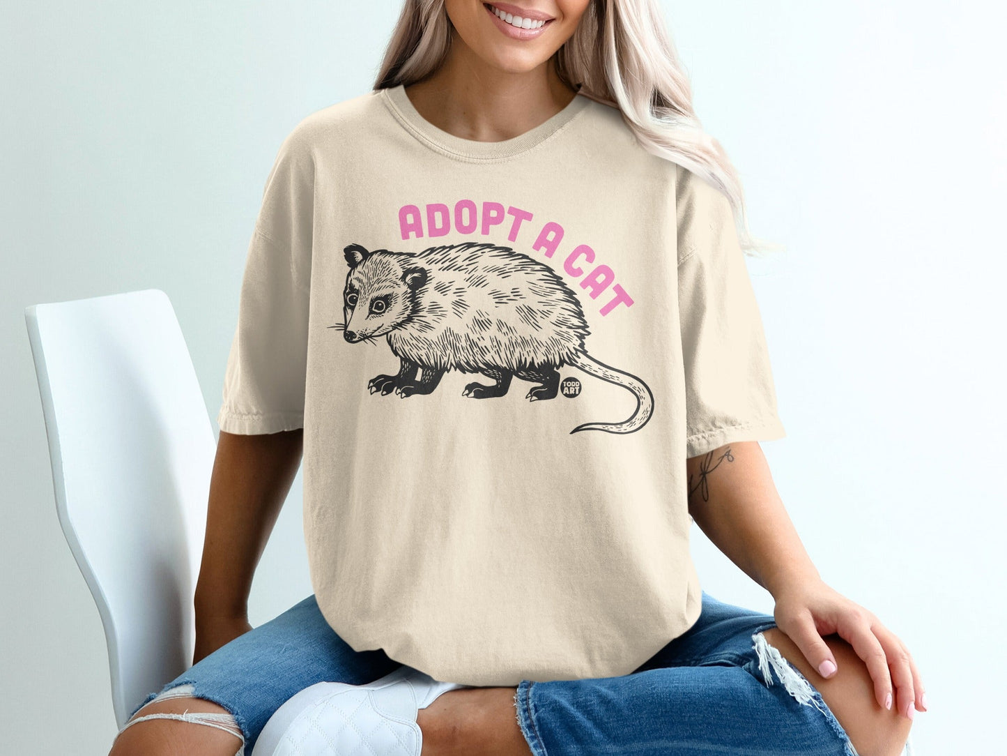 Opossum graphic tee with "ADOPT A CAT" text on cream-colored t-shirt.