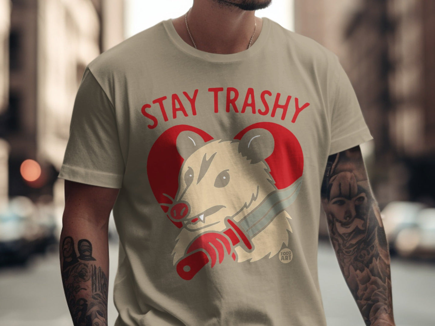 Tan t-shirt with red "STAY TRASHY" text and cartoon opossum holding a knife.
