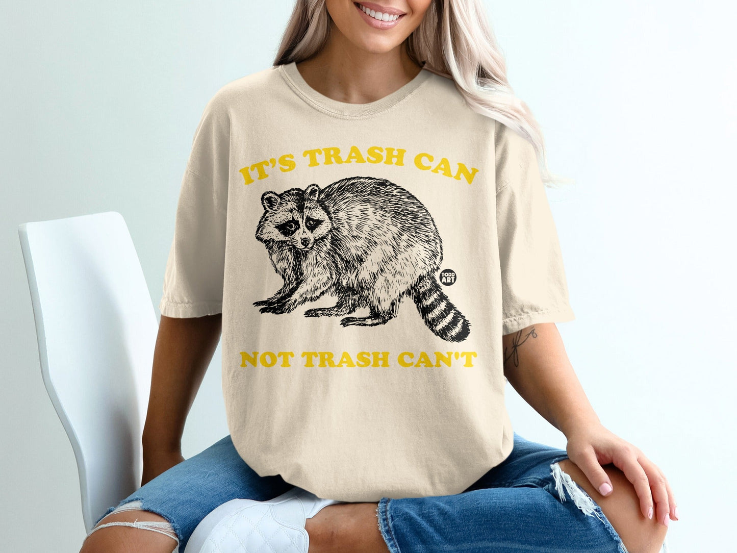 Cream t-shirt with raccoon graphic and yellow text "IT'S TRASH CAN NOT TRASH CAN'T".