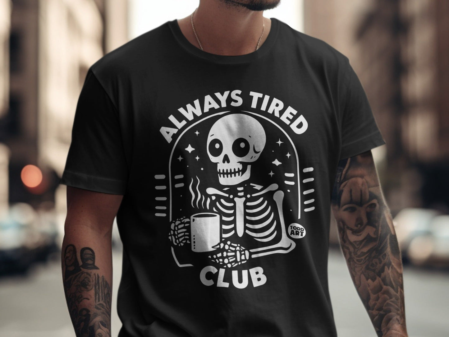 Black t-shirt with "Always Tired Club" skeleton holding coffee mug graphic.