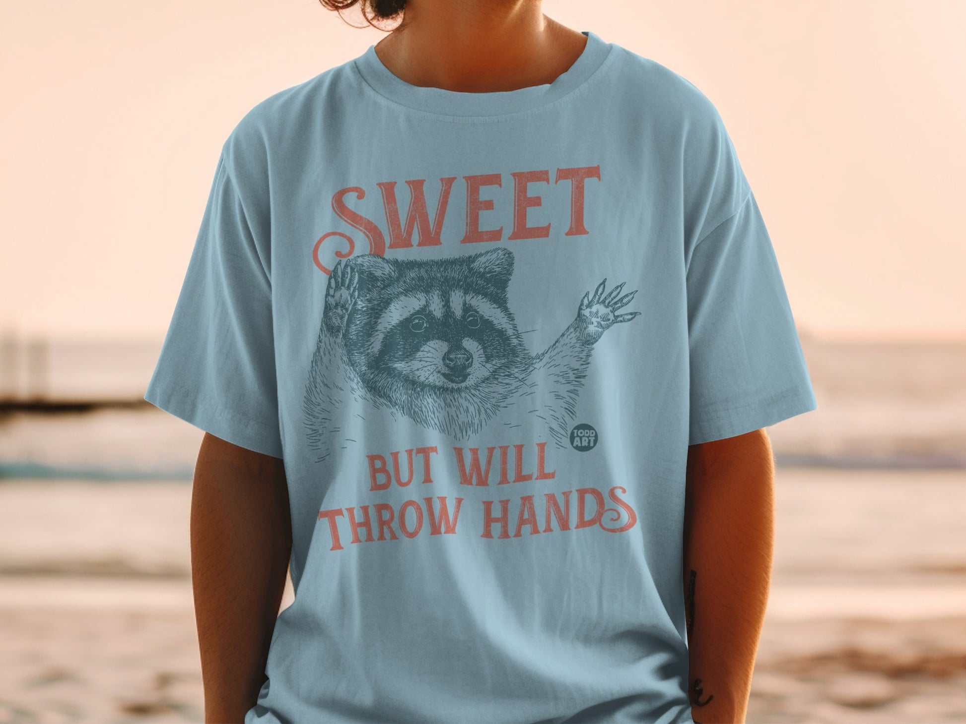 Light blue t-shirt with a raccoon illustration and text "SWEET BUT THROW HANDS".