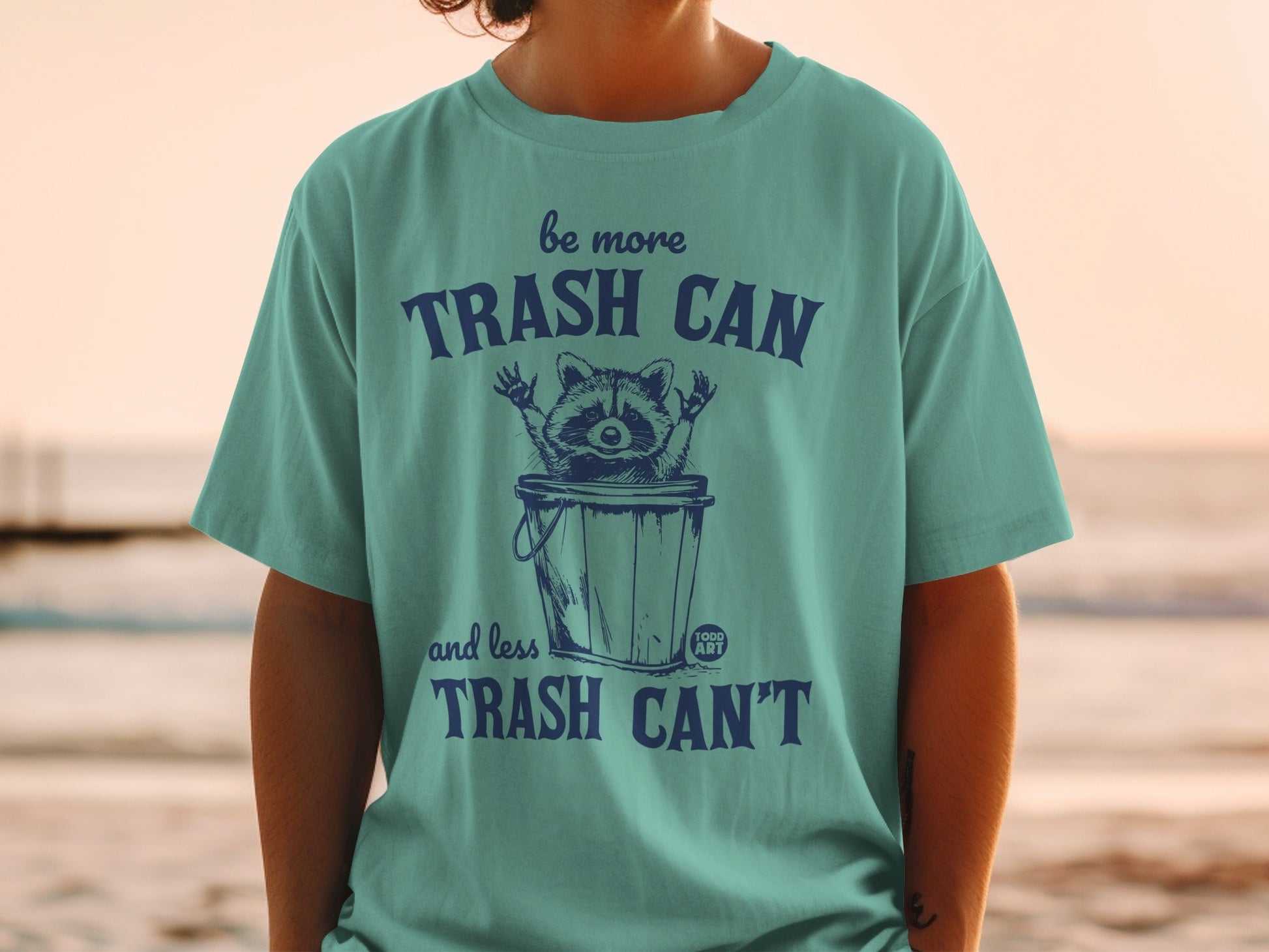 Teal t-shirt with raccoon in trash can and text "Be more trash can and less trash can't".