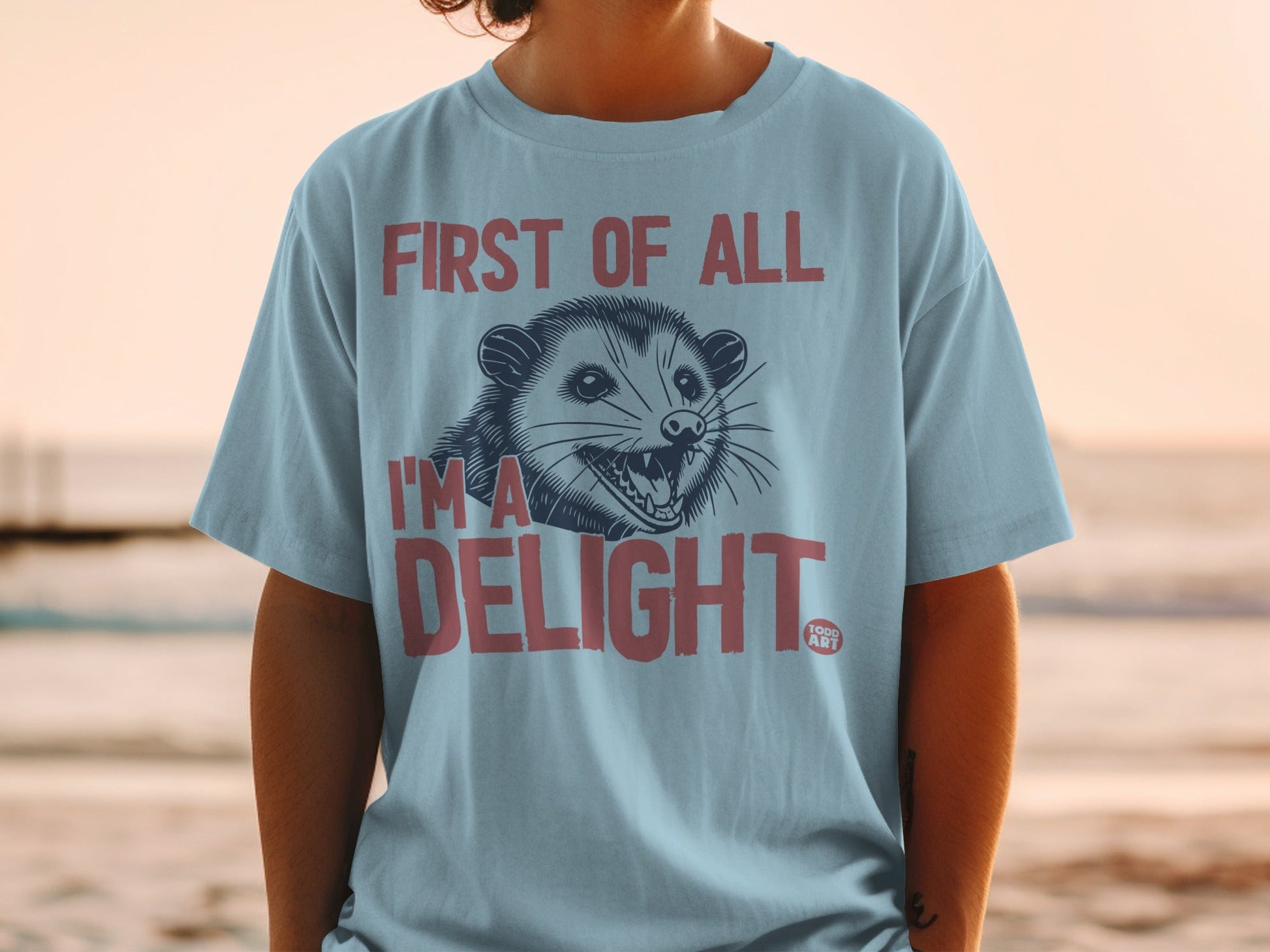 Light blue t-shirt with a grinning opossum and text "First of all I'm a DELIGHT".