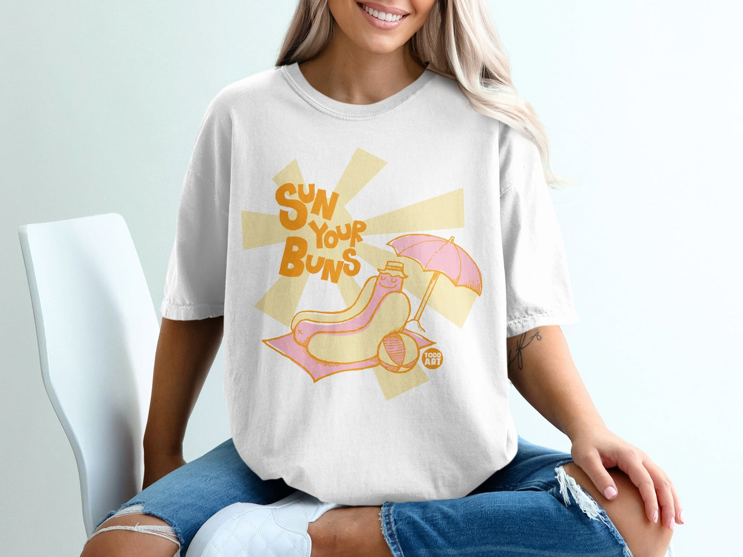 White t-shirt with cartoon hot dog on beach towel, text "Sun Your Buns".