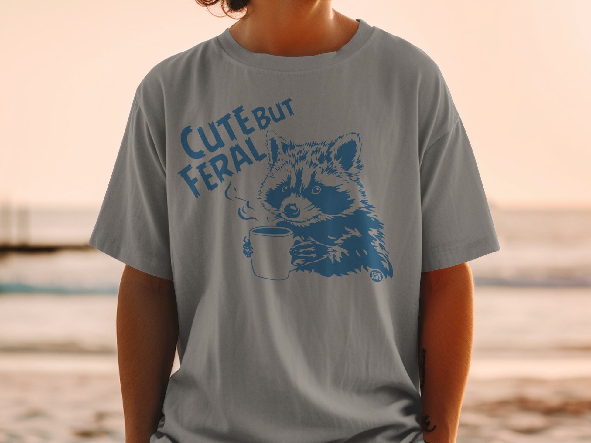 Gray t-shirt with blue graphic of a raccoon drinking coffee, "CUTE BUT FERAL" text.