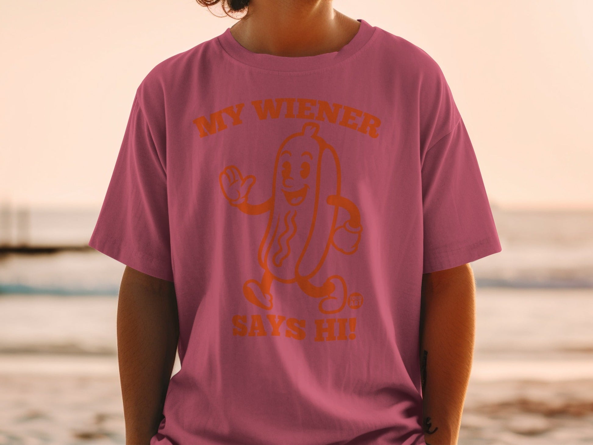 Orange graphic of a smiling hot dog with "MY WIENER SAYS HI!" on a pink t-shirt.