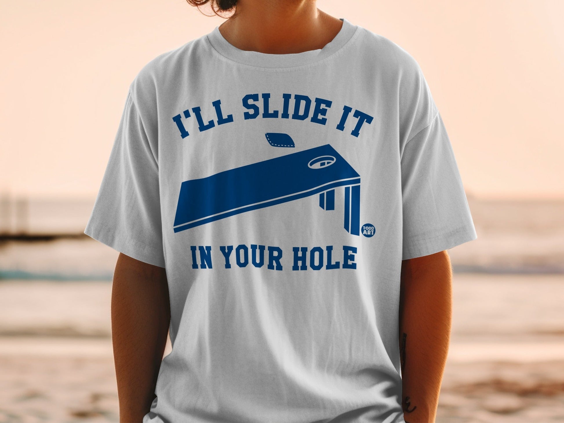 White t-shirt with dark blue graphic of a cornhole board and text.