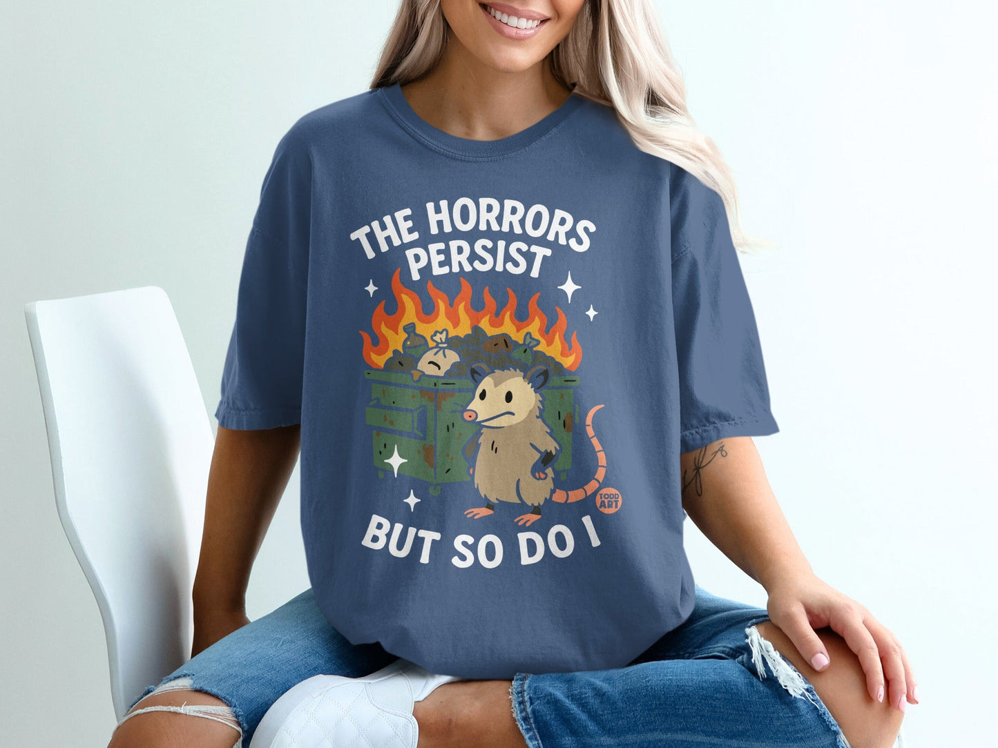 T-shirt with opossum in burning dumpster graphic and text "The horrors persist but so do I".