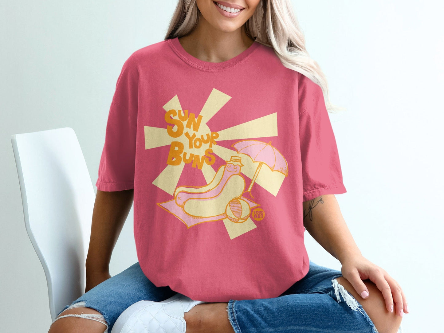 Pink t-shirt with graphic of a hot dog on a beach towel under an umbrella.