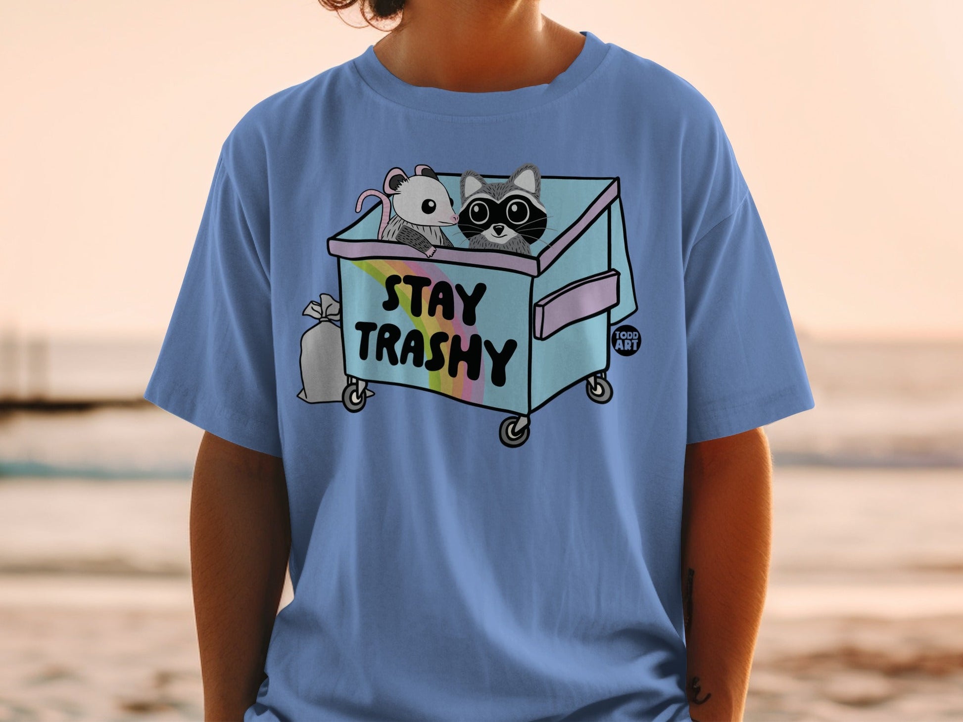 Blue t-shirt with raccoon and opossum in trash can graphic that says "STAY TRASHY".