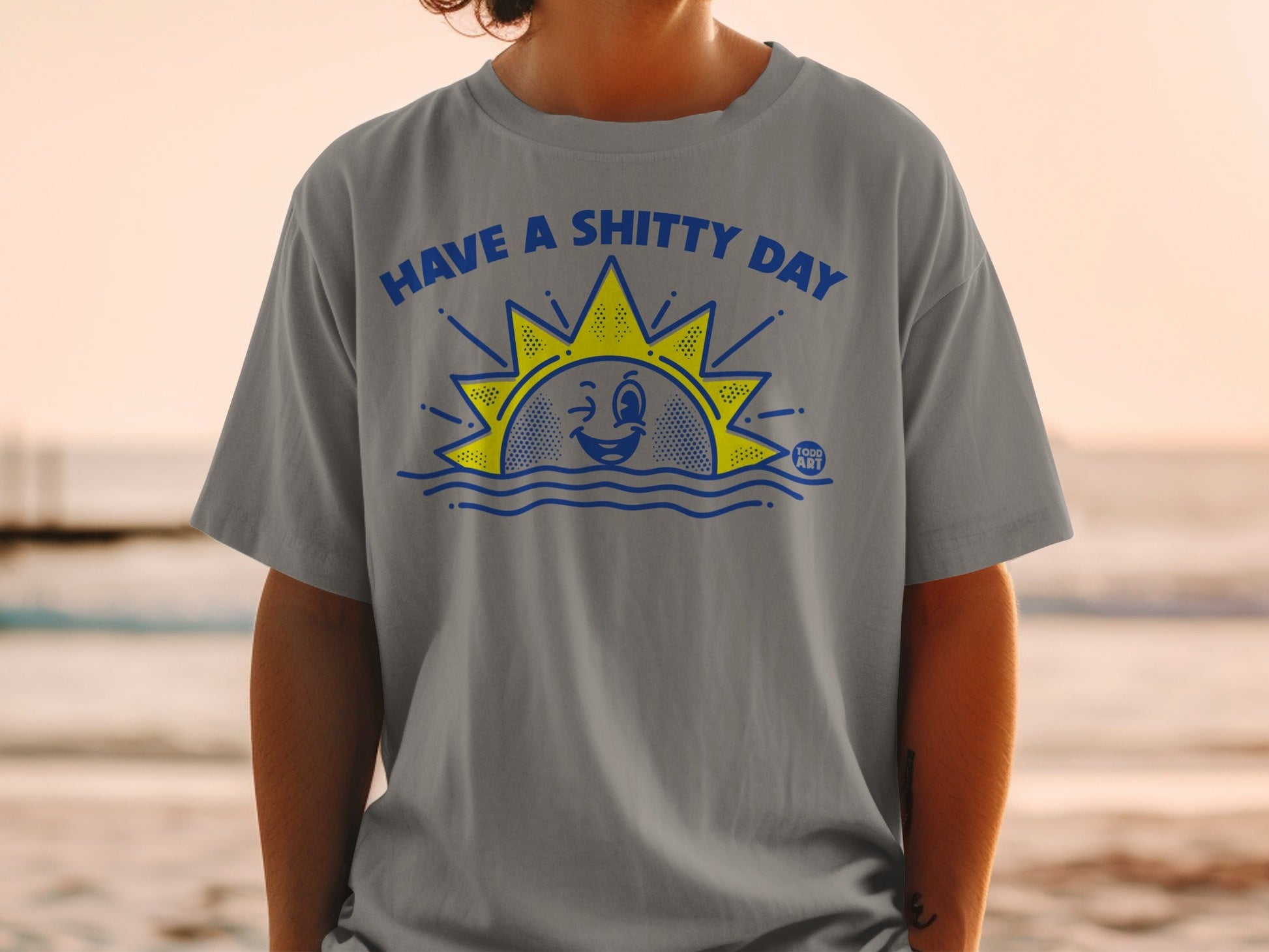 T-shirt with blue sun, waves, and text "HAVE A SHITTY DAY".