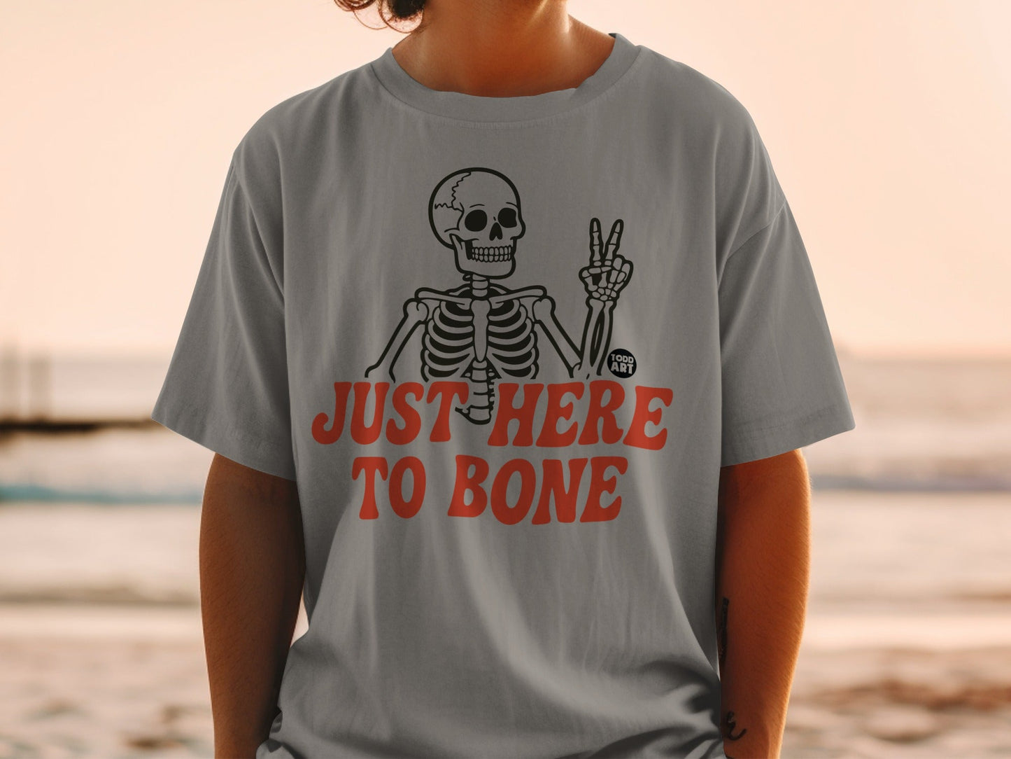 Gray t-shirt with skeleton giving peace sign and text "JUST HERE TO BONE".