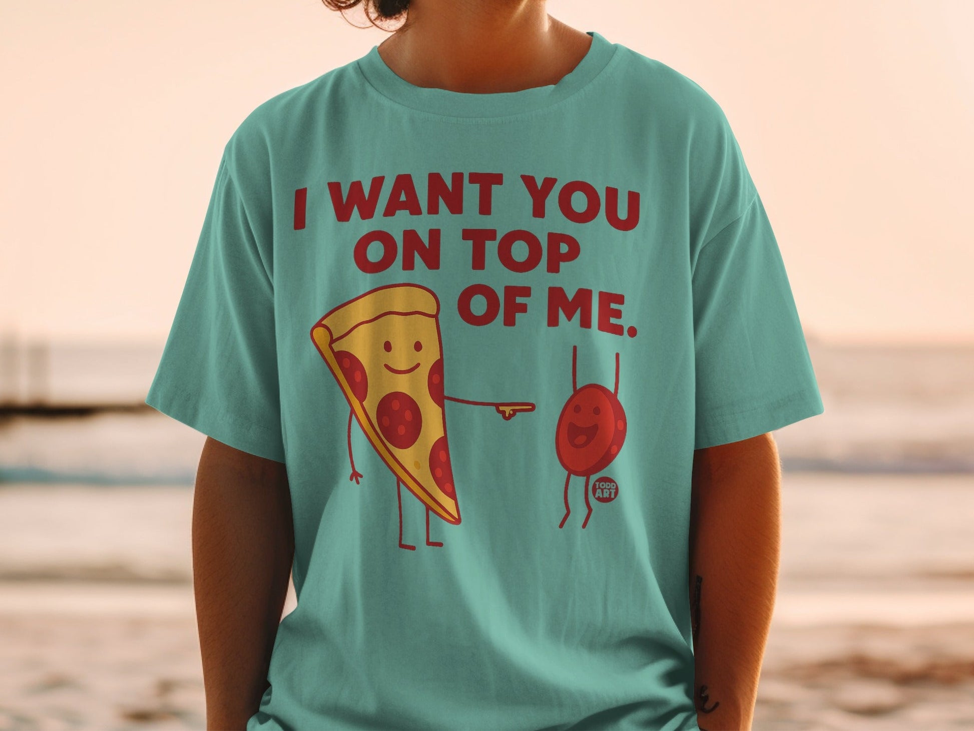 Teal t-shirt with text "I WANT YOU ON TOP OF ME" and pizza and balloon characters.