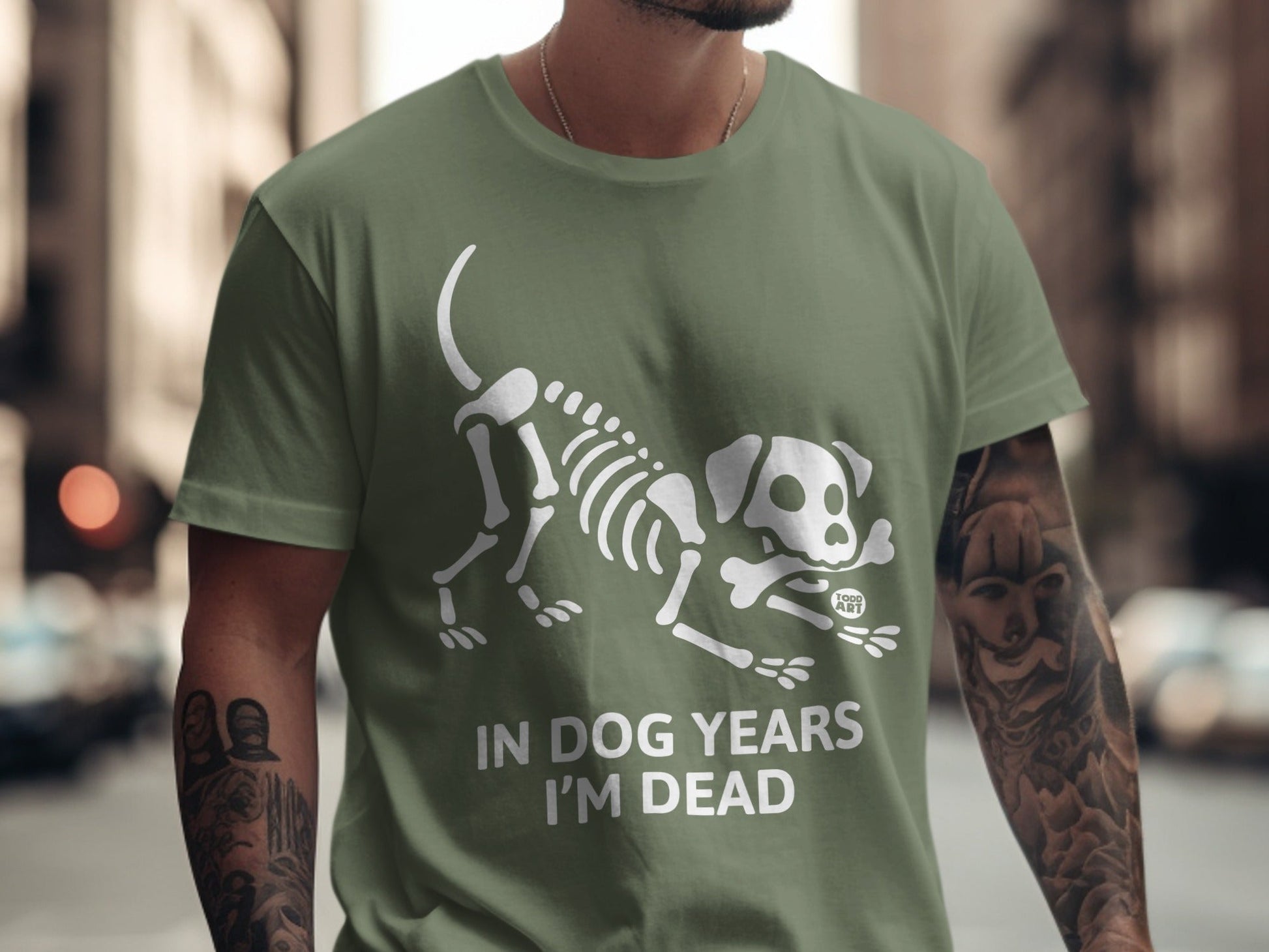Olive green t-shirt with white skeleton dog graphic and text "IN DOG YEARS I'M DEAD".