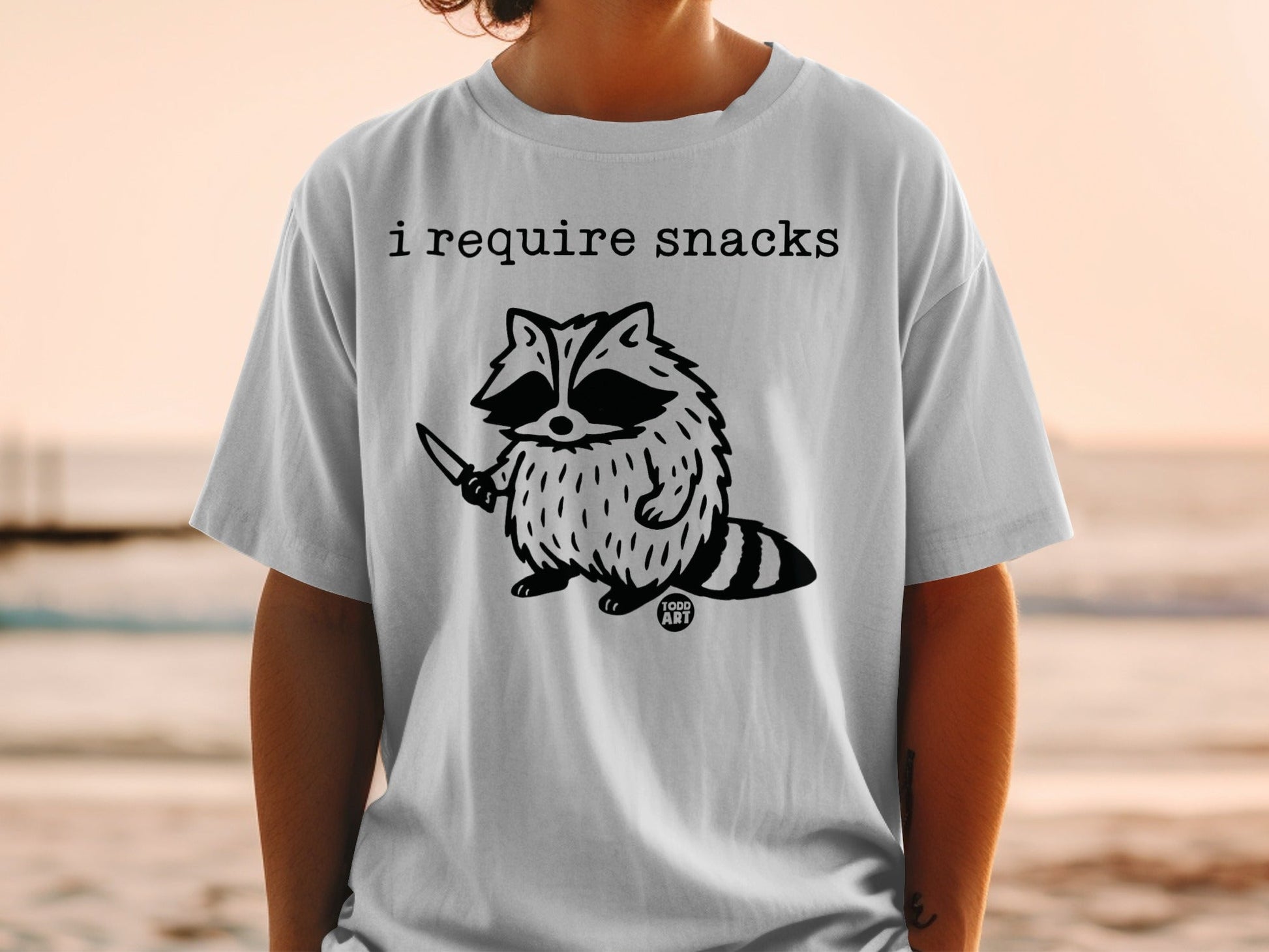 T-shirt with raccoon holding a knife and text "I REQUIRE SNACKS".
