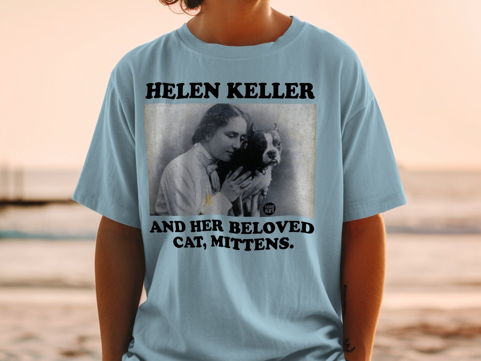 Light blue t-shirt with Helen Keller and her cat graphic and text.