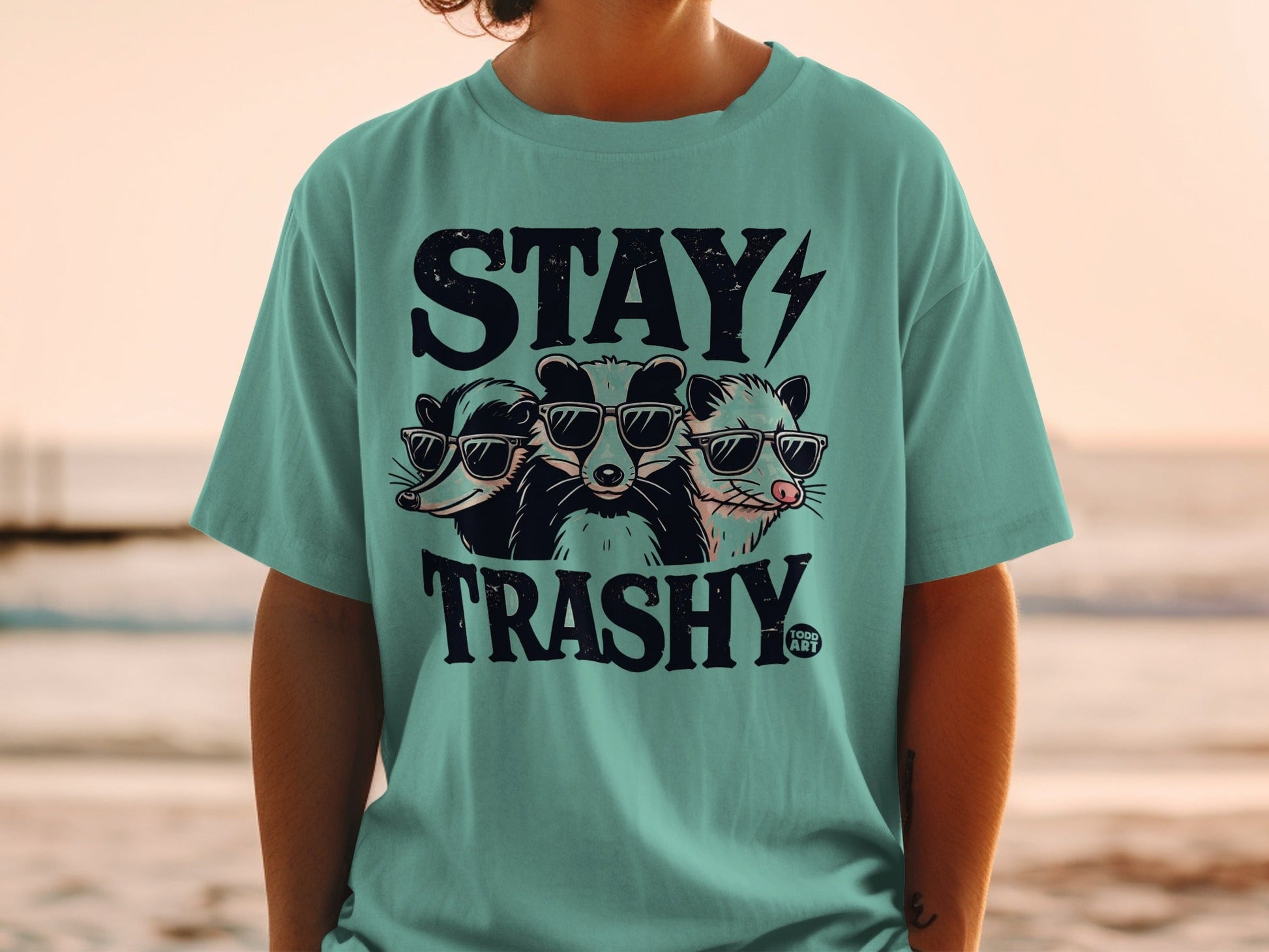 Three raccoons in sunglasses on a teal t-shirt with "STAY TRASHY" text.