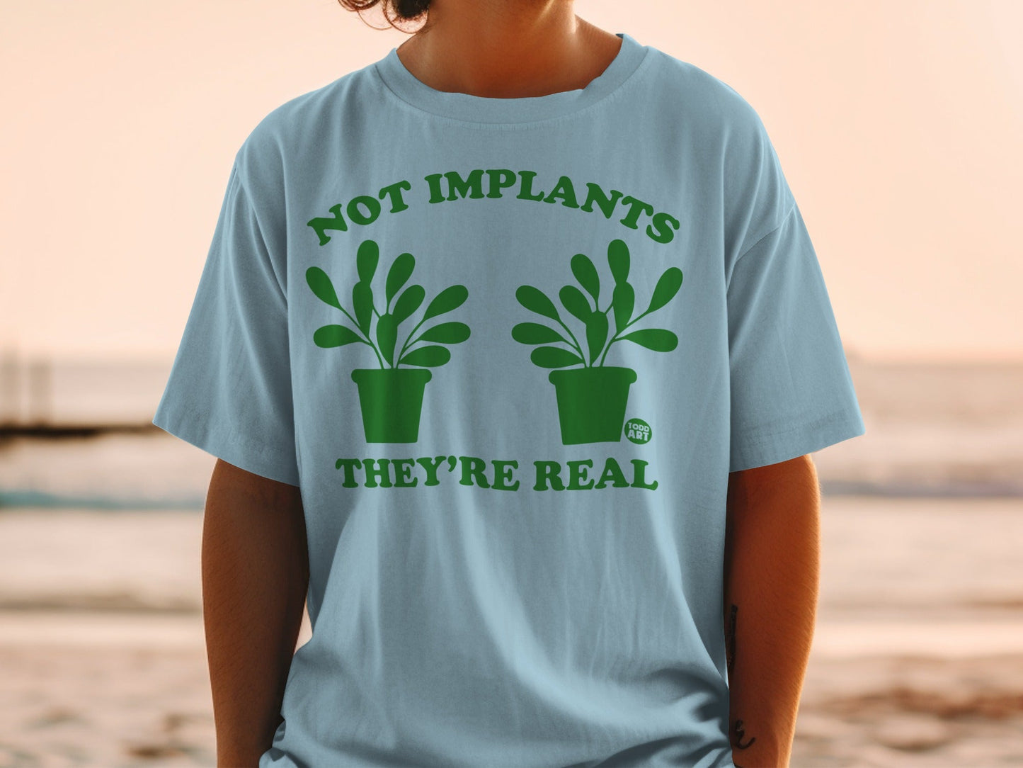Light blue t-shirt with green graphic reading "Not Implants They're Real" and two potted plants.