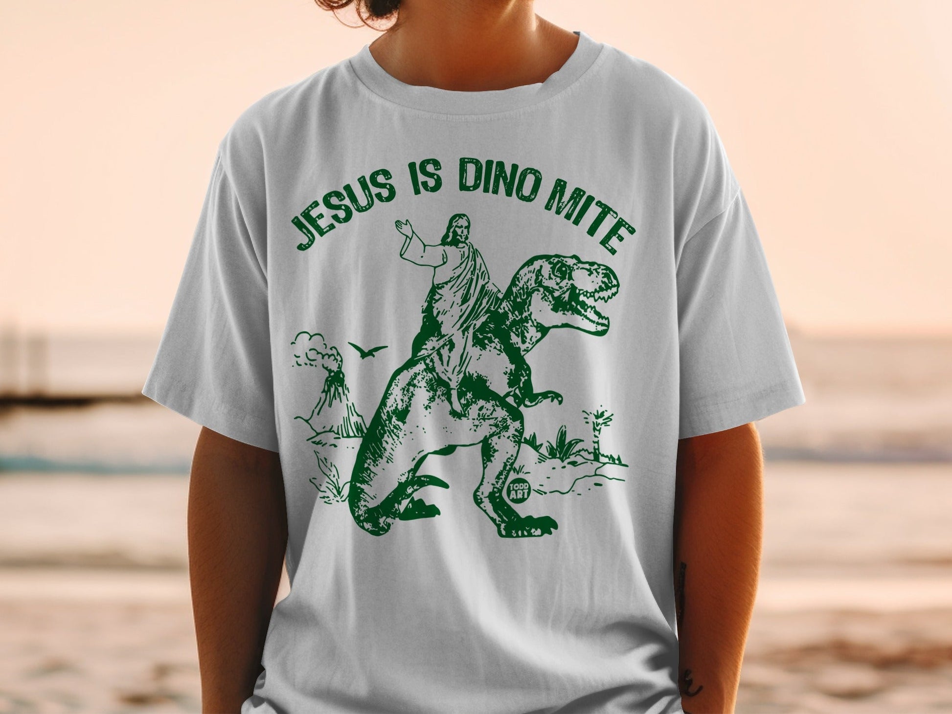 Jesus riding a Tyrannosaurus Rex with volcano background on a green t-shirt.