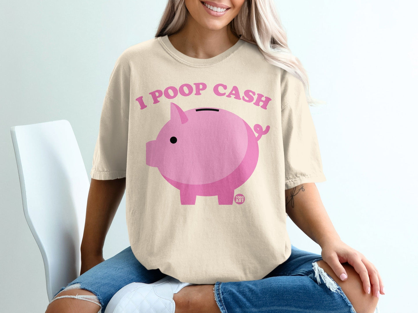 Pink piggy bank with text "I POOP CASH" on a light beige t-shirt.