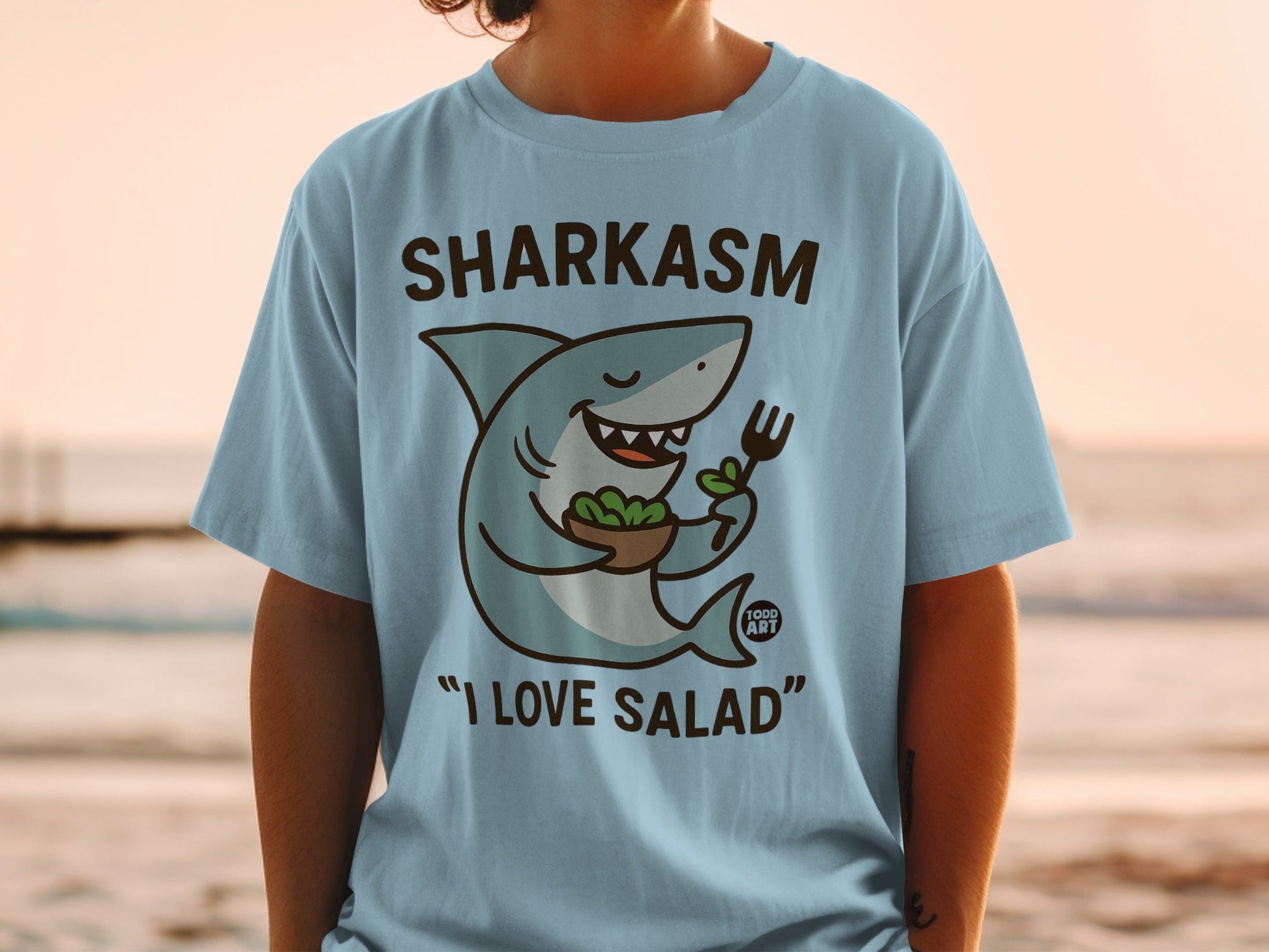 Light blue t-shirt with cartoon shark holding salad and fork, text "SHARKASM I LOVE SALAD".