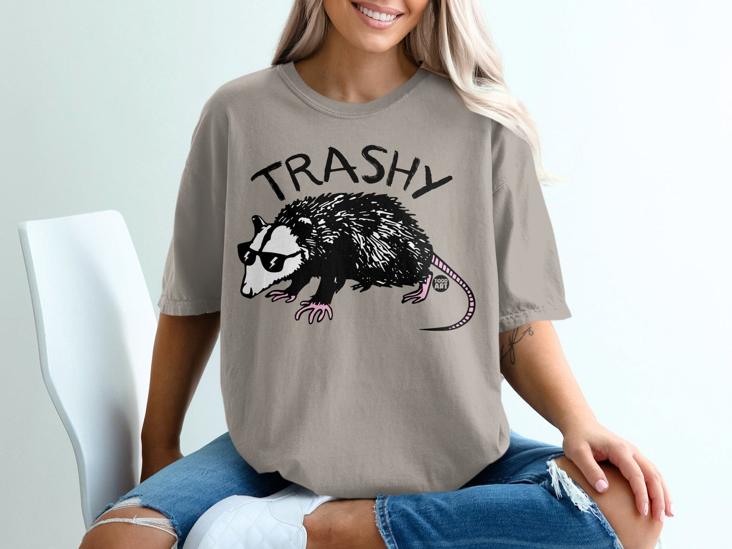 Opossum wearing sunglasses and a t-shirt with "TRASHY" printed on it.