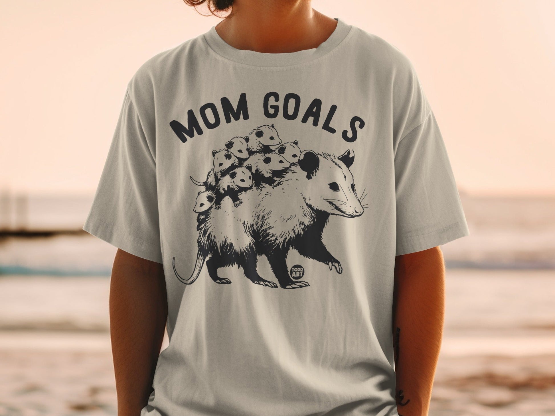 T-shirt with illustration of opossum and babies, text "MOM GOALS".