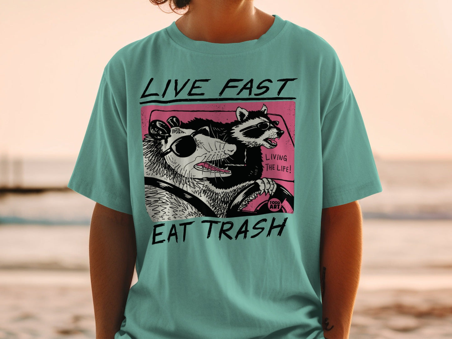Opossum and raccoon wearing sunglasses drive with "LIVE FAST EAT TRASH" text.