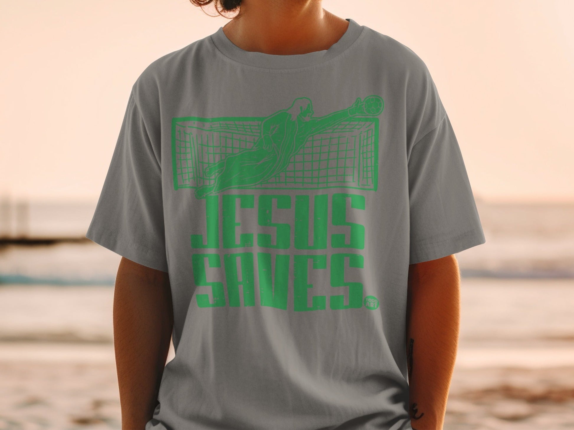 Green text "Jesus Saves" graphic on a gray t-shirt featuring a soccer goalkeeper.