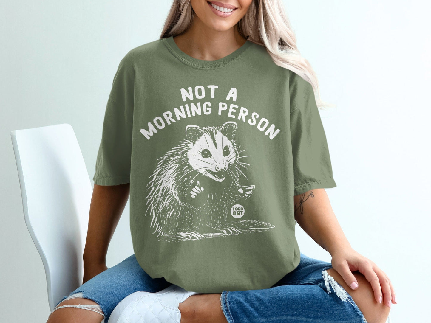 Olive green t-shirt with white text "NOT A MORNING PERSON" and a cartoon opossum.