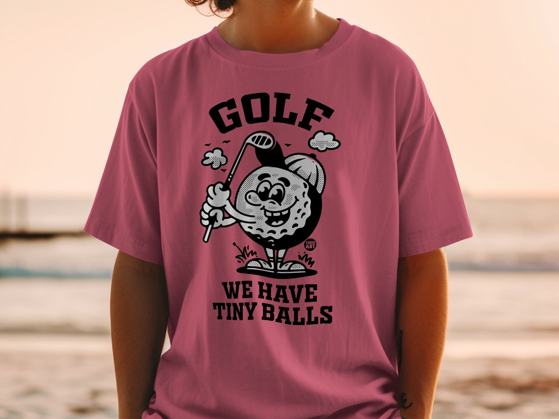 Pink t-shirt with cartoon golf ball character and text "GOLF WE HAVE TINY BALLS".