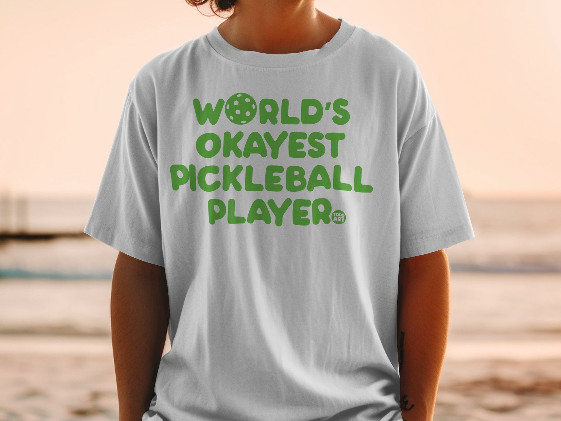 White t-shirt with green text "World's Okayest Pickleball Player" and a pickleball logo.
