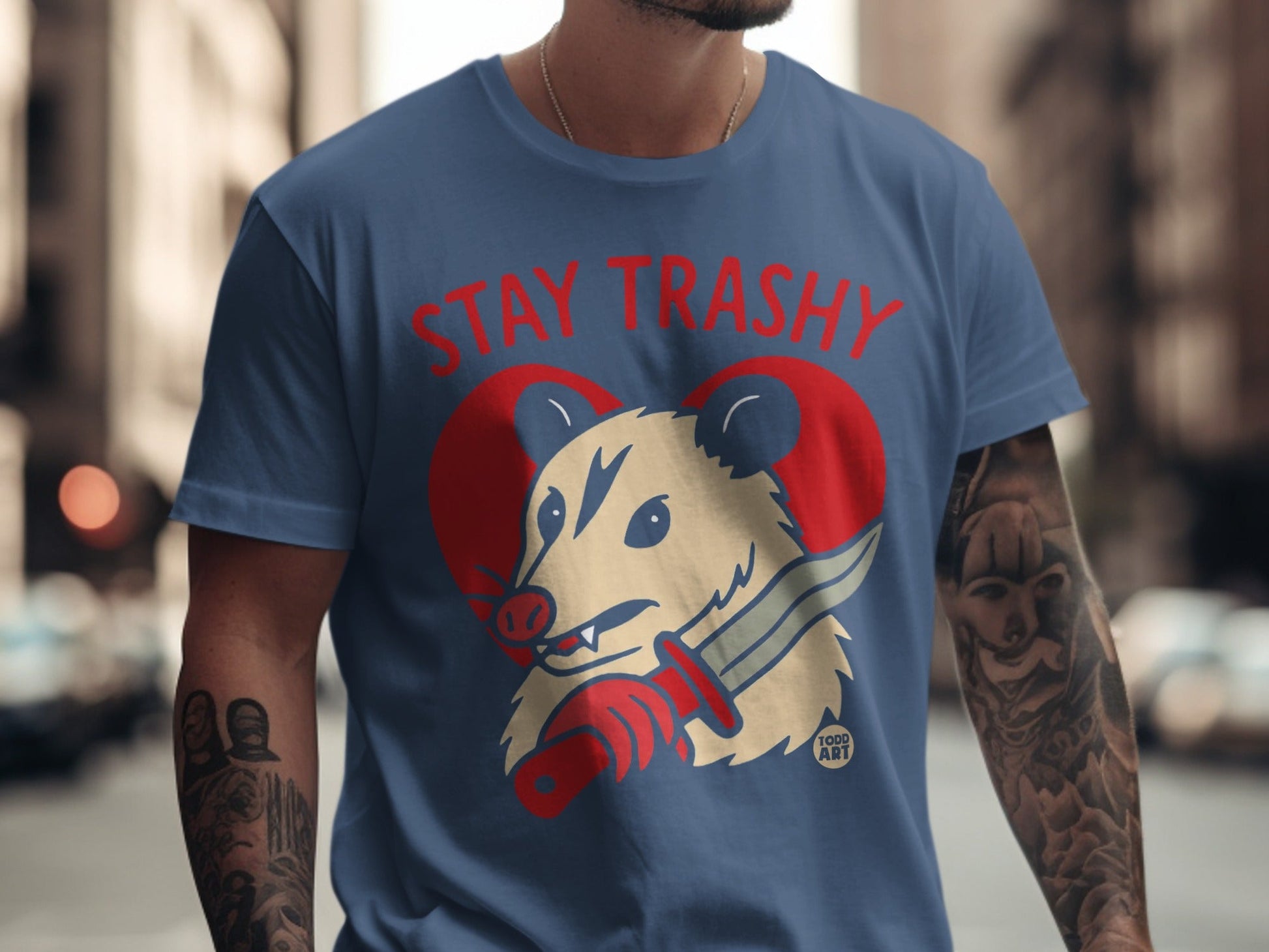 Blue t-shirt with a possum wearing a red heart and holding a knife, with "STAY TRASHY" text.
