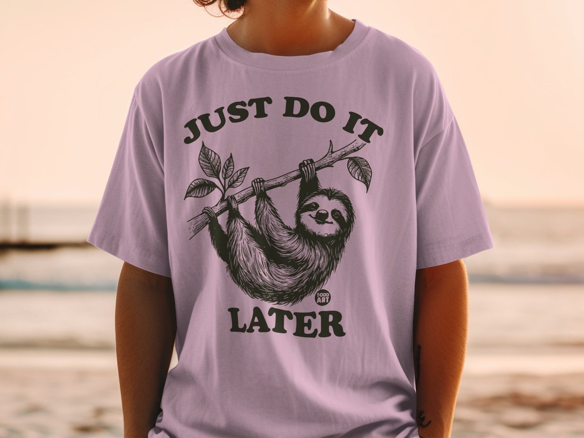 Sloth hanging from branch on purple t-shirt, text reads "JUST DO IT LATER".