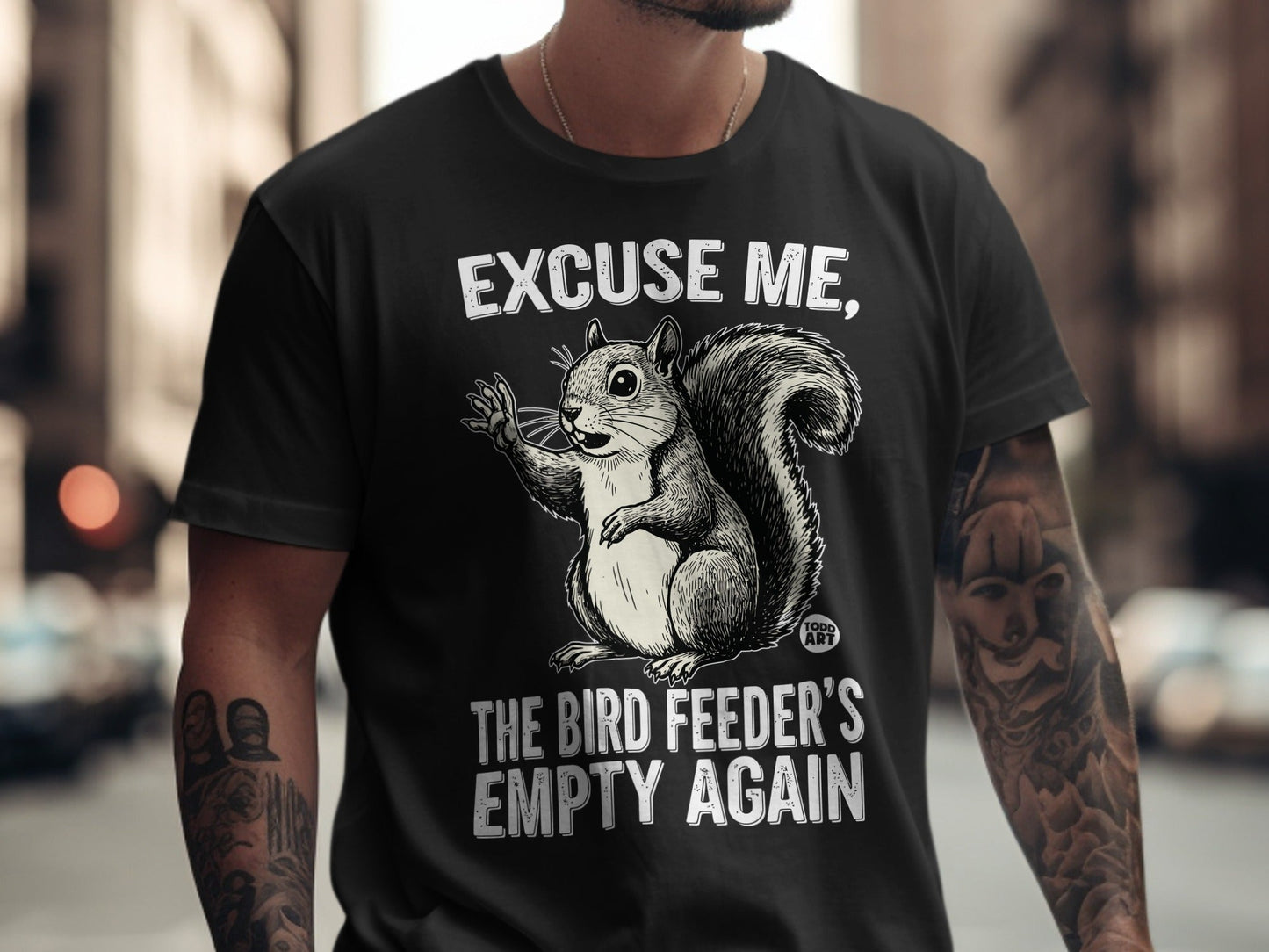 Black t-shirt with a squirrel illustration and text: "Excuse me, the bird feeder's empty again".