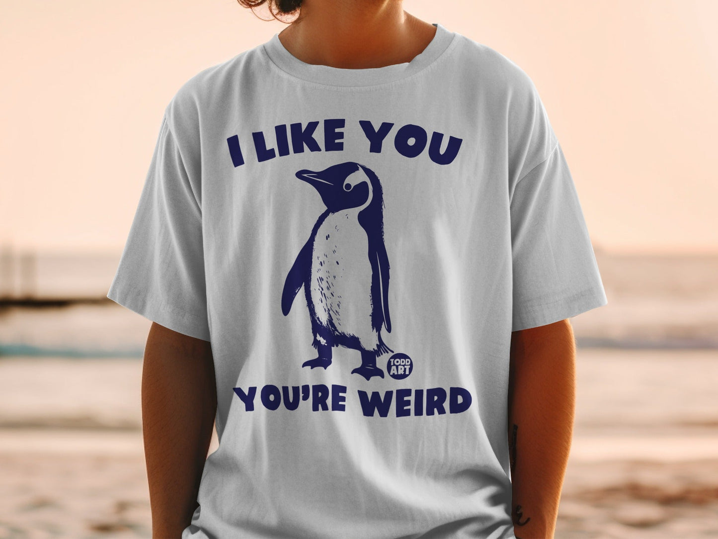 White t-shirt with a penguin graphic and text "I LIKE YOU YOU'RE WEIRD".