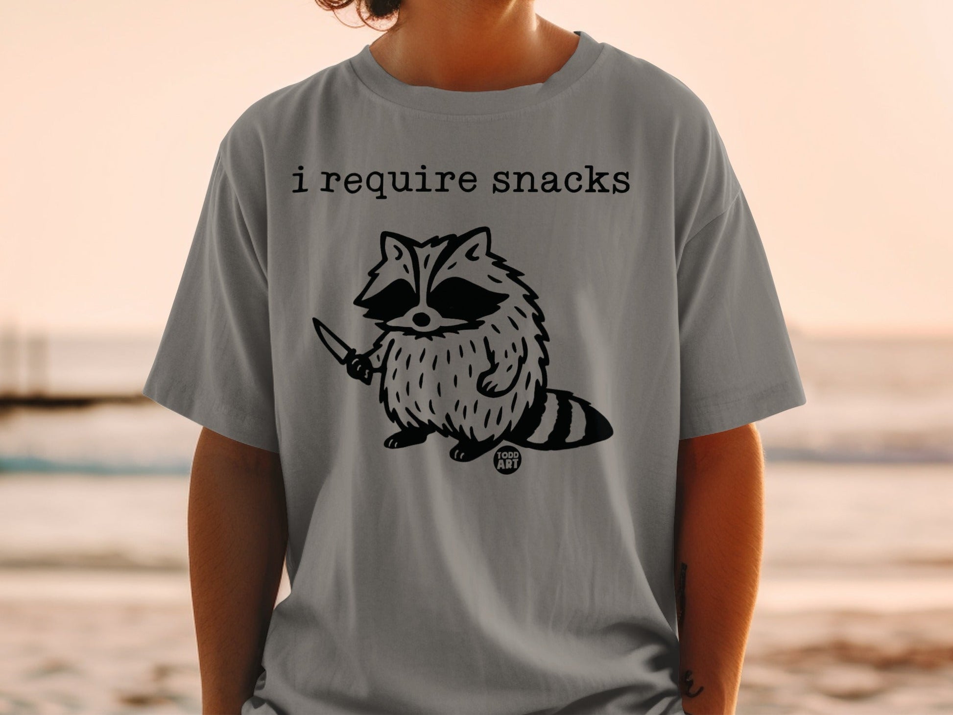 T-shirt with a raccoon holding a knife and the text "I require snacks."
