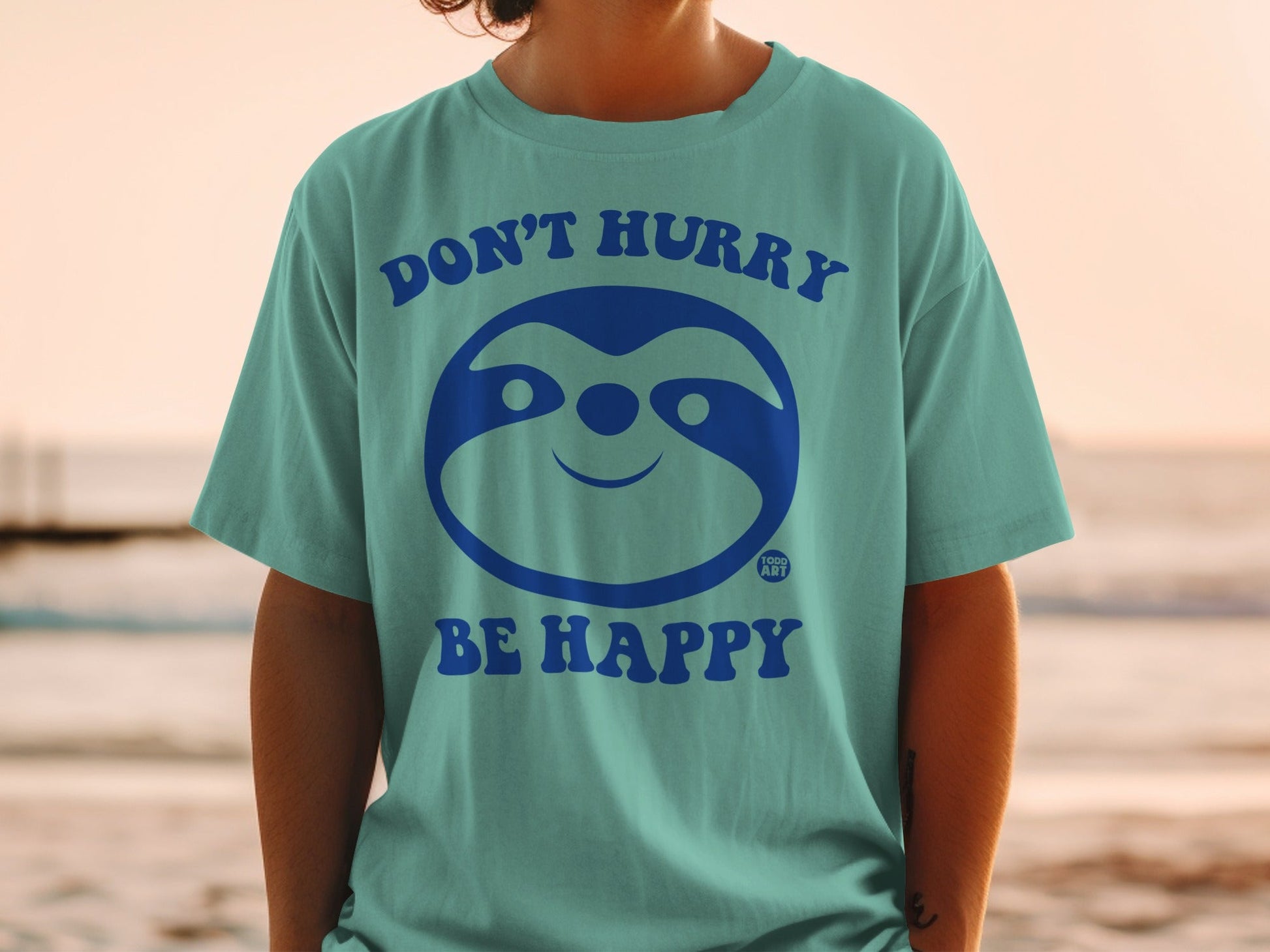 Teal t-shirt with a smiling sloth graphic and text "DON'T HURRY BE HAPPY".