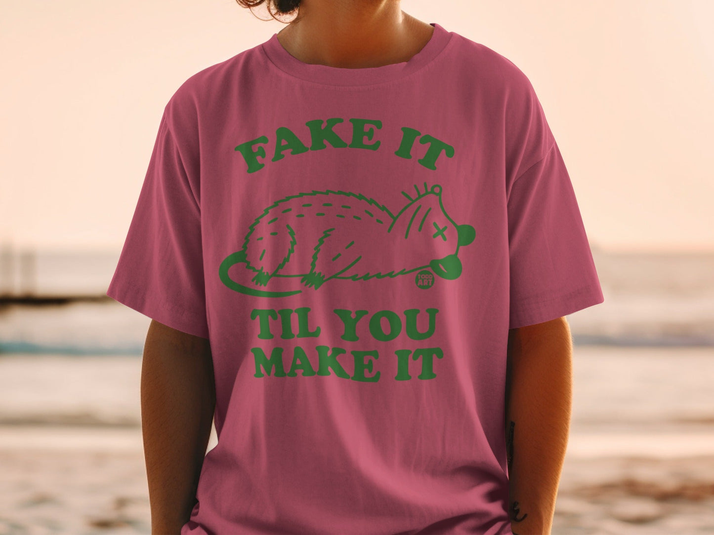 Pink t-shirt with green text and possum graphic, "Fake It Til You Make It".