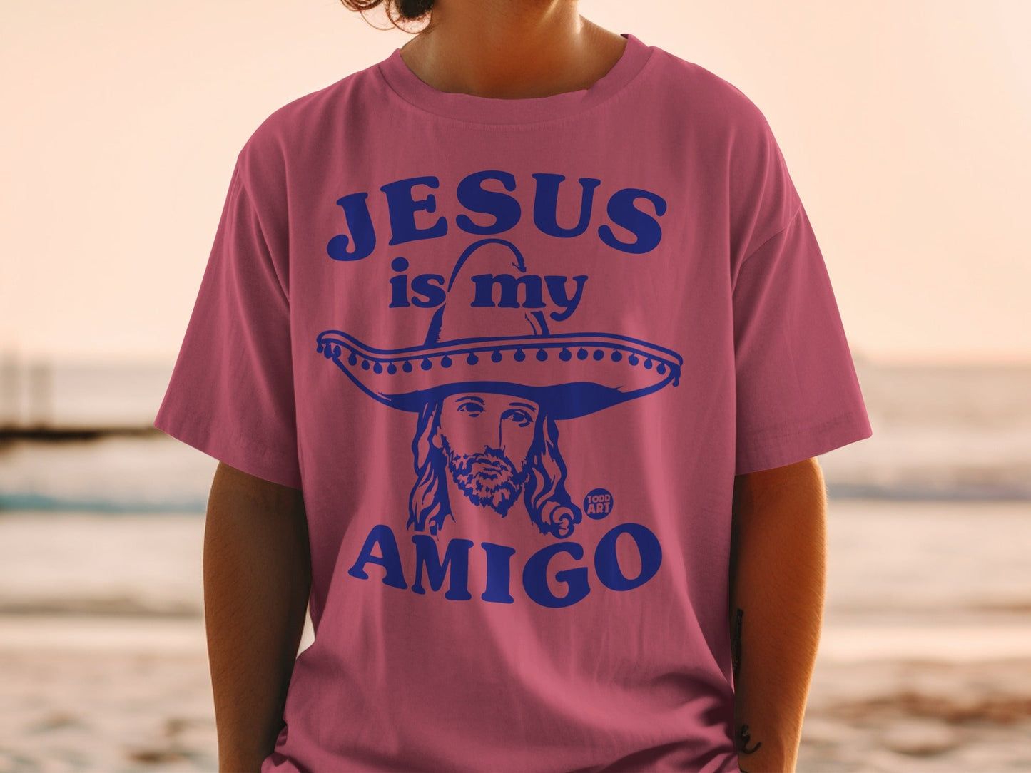 Pink t-shirt with blue text reading "Jesus is my Amigo" and a sombrero-wearing Jesus graphic.