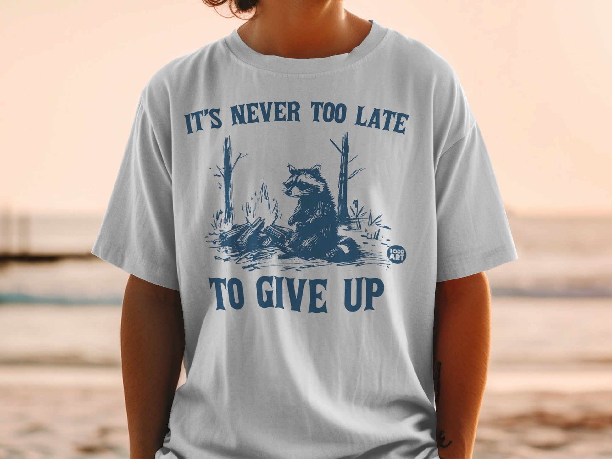 T-shirt graphic with raccoon by campfire, text "IT'S NEVER TOO LATE TO GIVE UP".