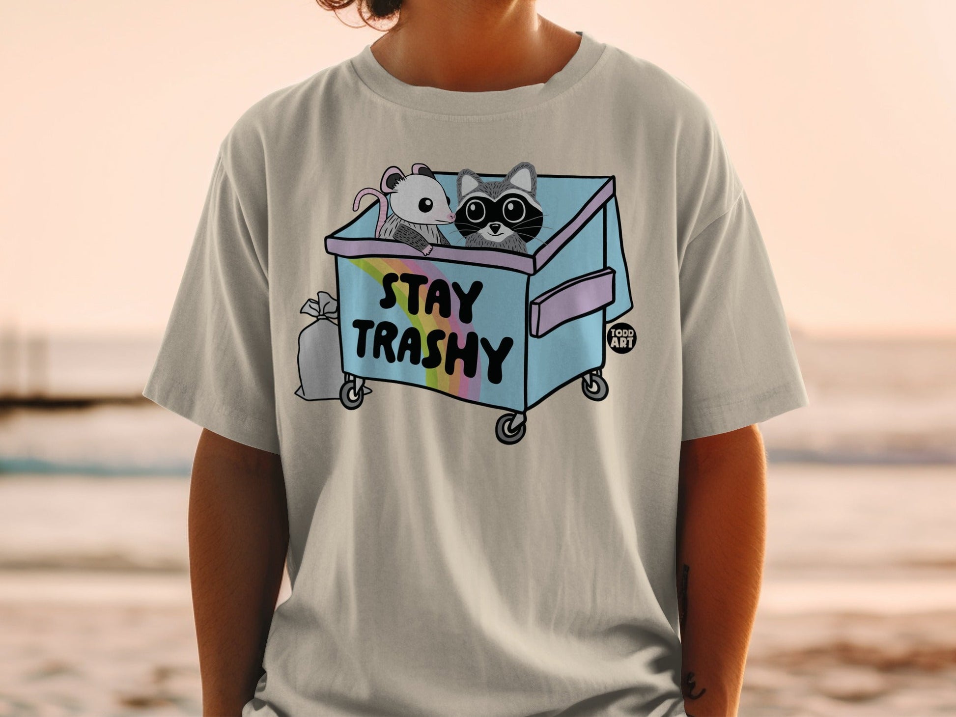 T-shirt graphic of a raccoon and mouse in a trash can with "STAY TRASHY" text.