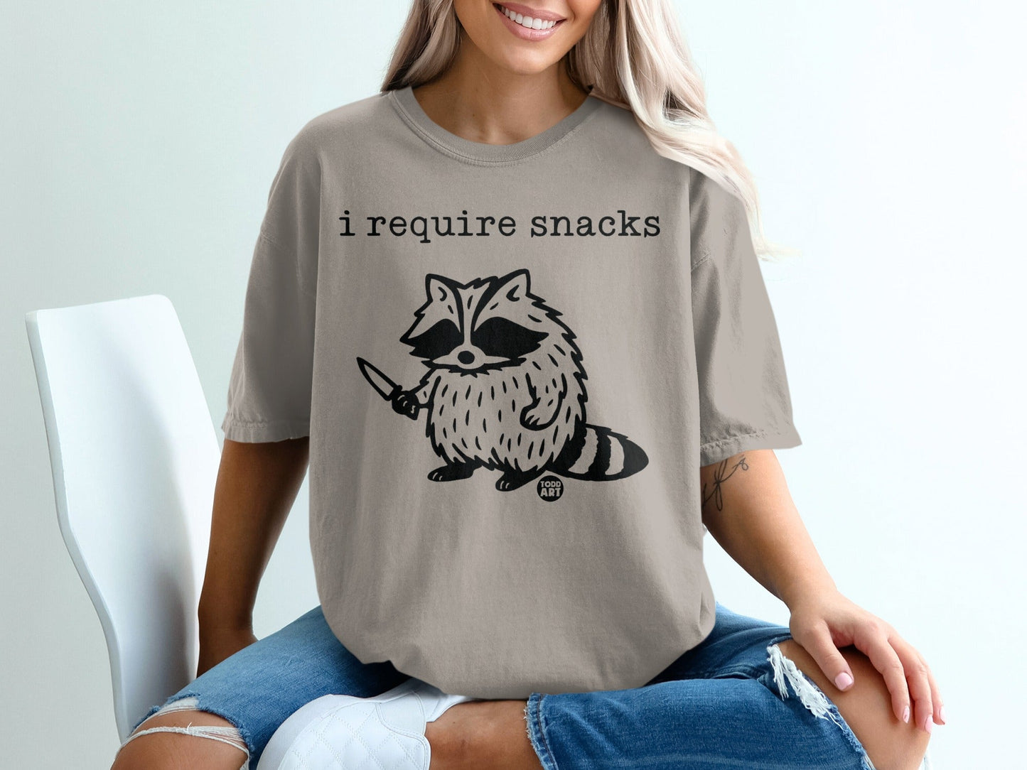 Tan t-shirt with raccoon graphic and text "i require snacks".