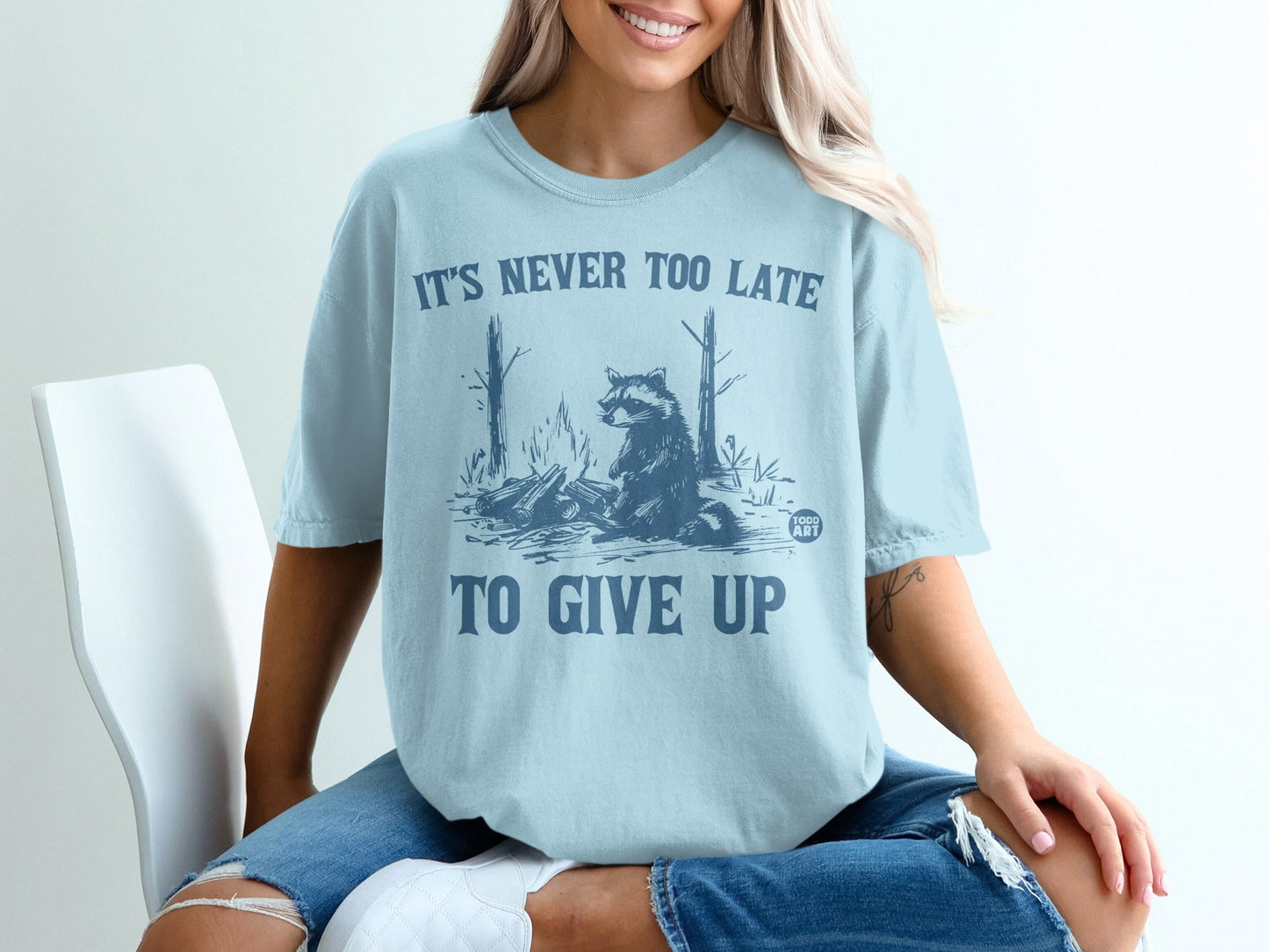 Light blue t-shirt with raccoon by campfire graphic and text "It's never too late to give up."