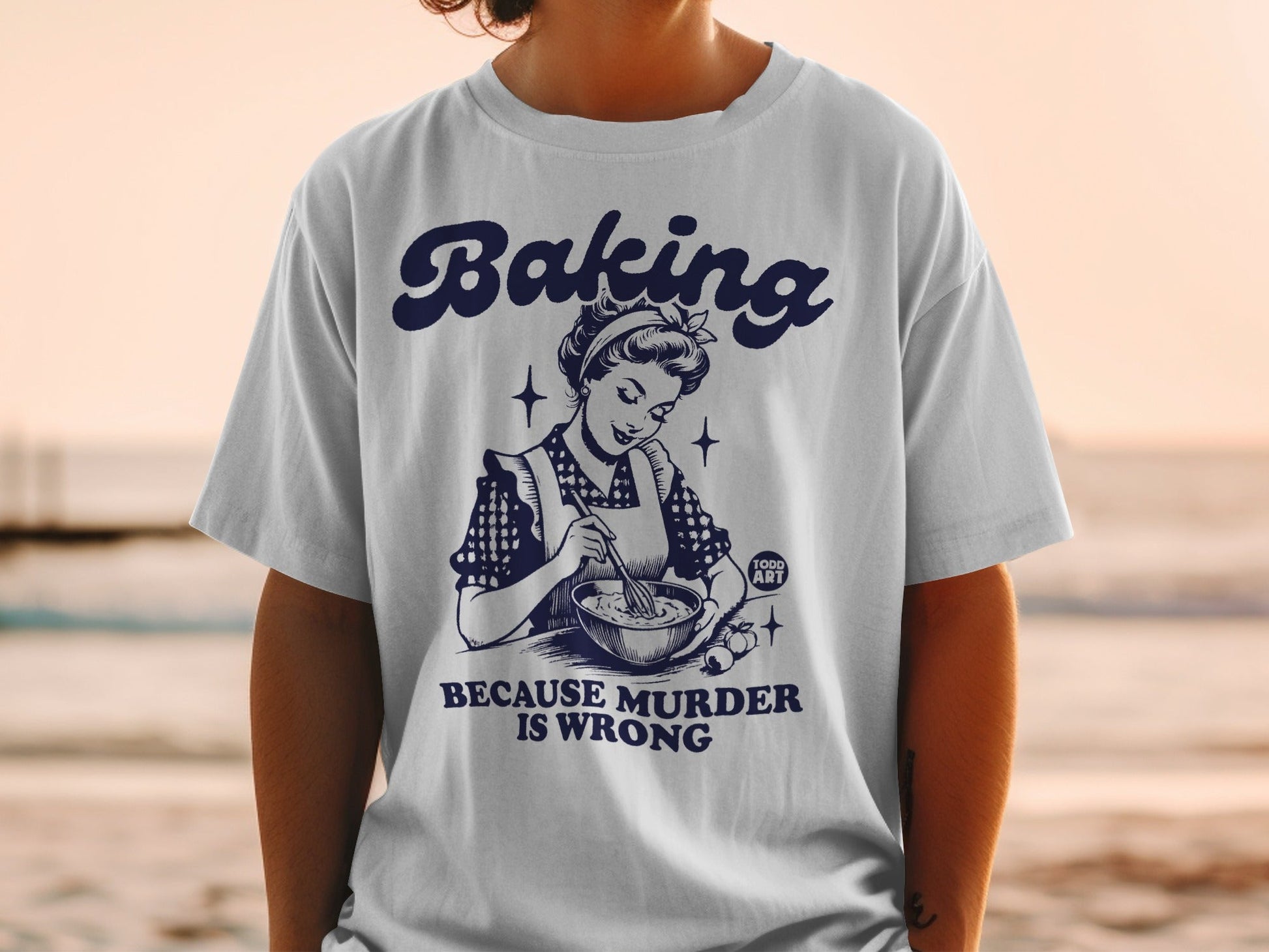 White t-shirt with "Baking Because Murder Is Wrong" text and retro illustration.