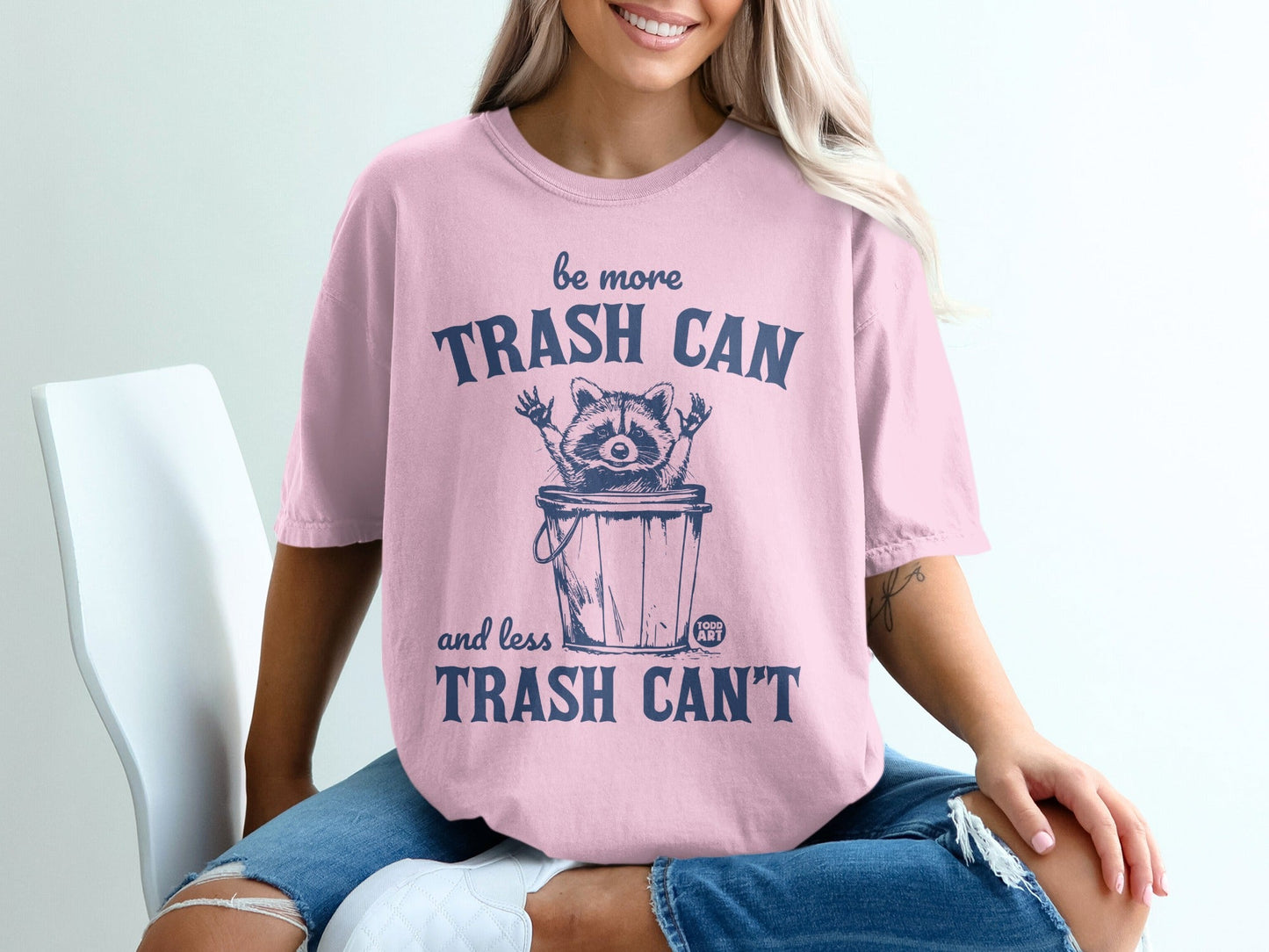Pink t-shirt with raccoon in trash can graphic and "be more trash can and less trash can't" text.