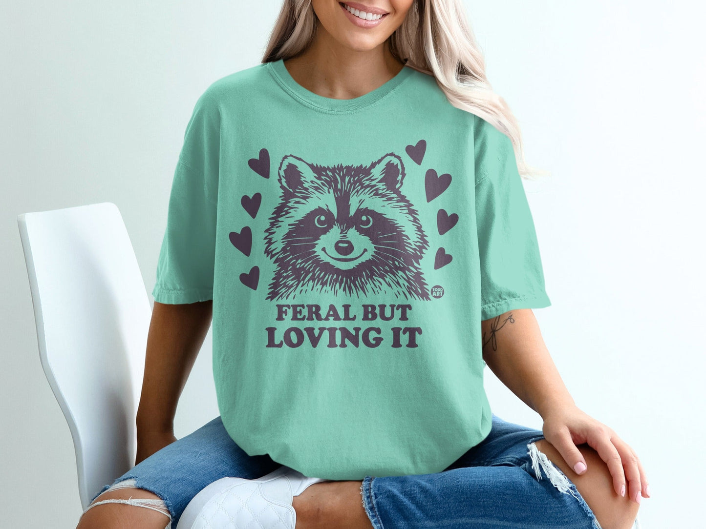 Mint t-shirt with raccoon graphic and "Feral but loving it" text.