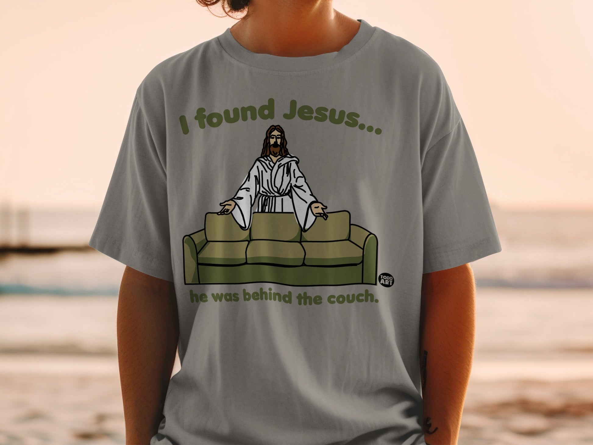 Grey t-shirt with "I found Jesus... he was behind the couch." text and illustration.
